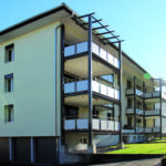 Ein modernes dreistöckiges Wohnhaus mit weißen Wänden und Balkonen auf jeder Etage. Das Gebäude verfügt über ein Flachdach und große Fenster. Das Gebäude ist von einer gepflegten Rasenfläche umgeben und der Himmel ist klar und sonnig.